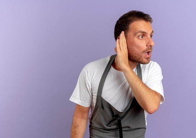 Free Photo barber man in apron holding hair cutting machine holding hand near his ear trying to listen standing over purple wall