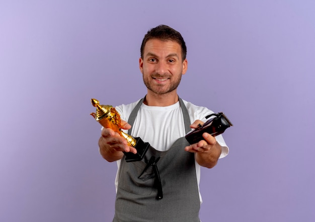 Barber man in apron holding hair cutting machine and trophy looking to the front smiling confident standing over purple wall