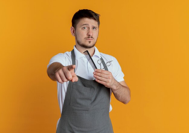 Barber man in apron holding scissors pointing with finger to camera with serious face standing over orange background