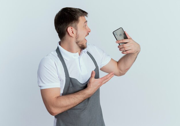 Barber man in apron holding smartphone looking at it excited standing over white background