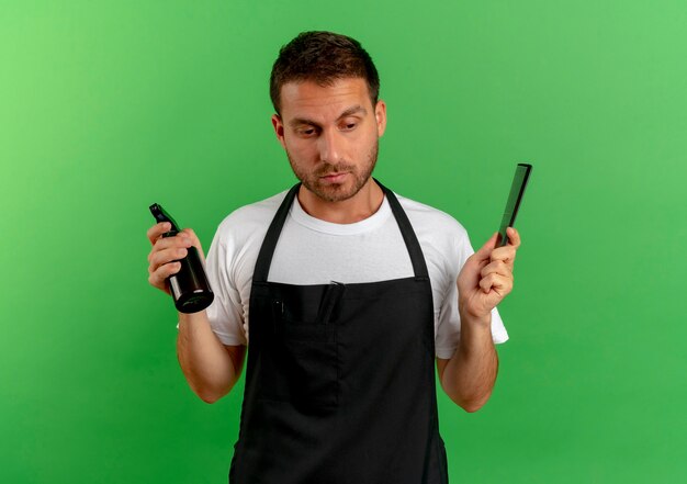 Barber man in apron holding spray and comb looking aside puzzled standing over green wall