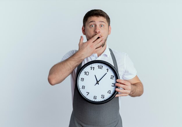 Barber man in apron holding wall clock looking at camera covering mouth with hand being shocked standing over white background