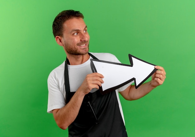 Free photo barber man in apron holding white arrow looking aside with smile on face standing over green wall