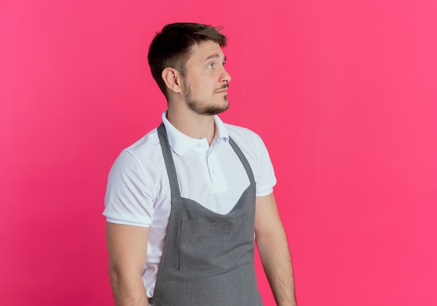 Barber man in apron looking aside with sad expression standing over pink background