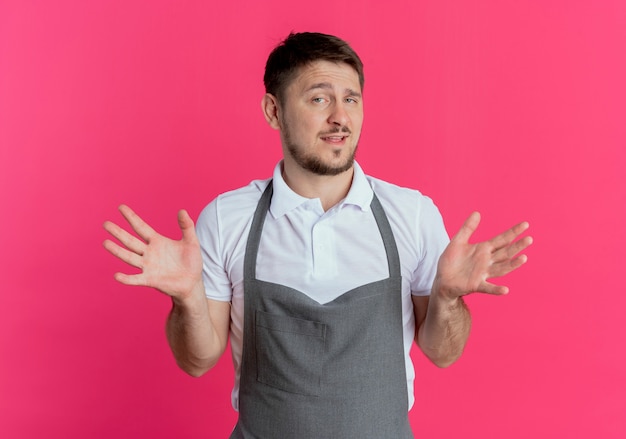 Barber man in apron looking at camera confused smiling spreading arms to the sides standing over pink background