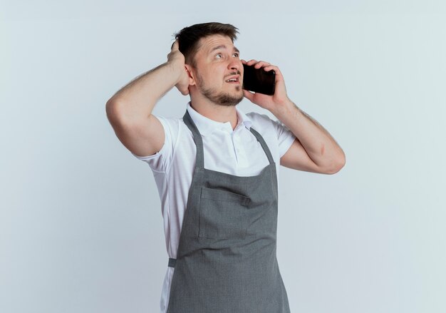 Barber man in apron looking confused while talking on mobile phone standing over white background