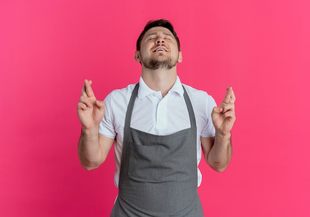 Free photo barber man in apron making desirable wish crossing fingers with eyes closed standing over pink wall