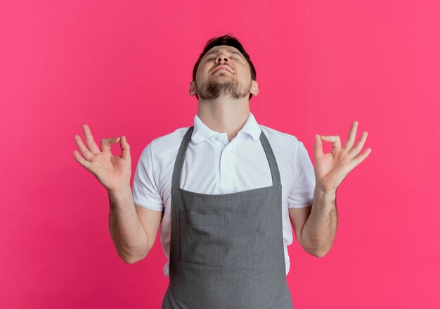 Barber man in apron relaxing making meditation gesture with fingers with eyes closed standing over pink background