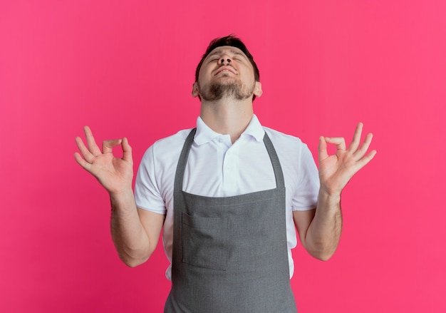 barber man in apron relaxing making meditation gesture with fingers with eyes closed standing over pink wall