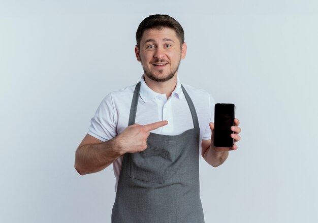 barber man in apron showing smartphone pointing with finger to it smiling standing over white wall