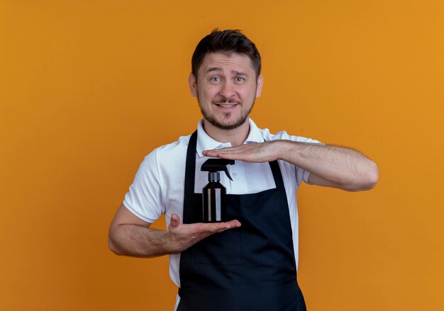 Barber man in apron showing spray looking at it with smile on face standing over orange background