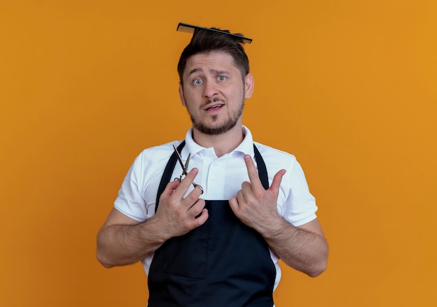 Barber man in apron with hair brush in his hair holding scissors looking at camera confused standing over orange background