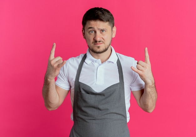 Free photo barber man in apron  with serious face showing rock symbol standing over pink wall