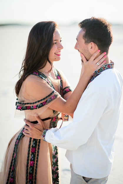 Free photo barefooted couple in bright embroidered clothing hugs tender on a white sand