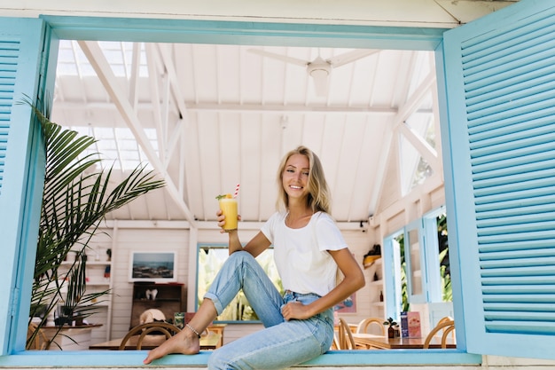 Barefooted girl drinking cocktail on window sill. Attractive young lady wears leg bracelet posing with amazing smile.