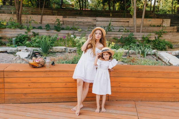 Barefooted slim woman and her daughter in white dress standing on wooden floor on nature. Good-looking shapely lady posing in park with little niece after picnic.