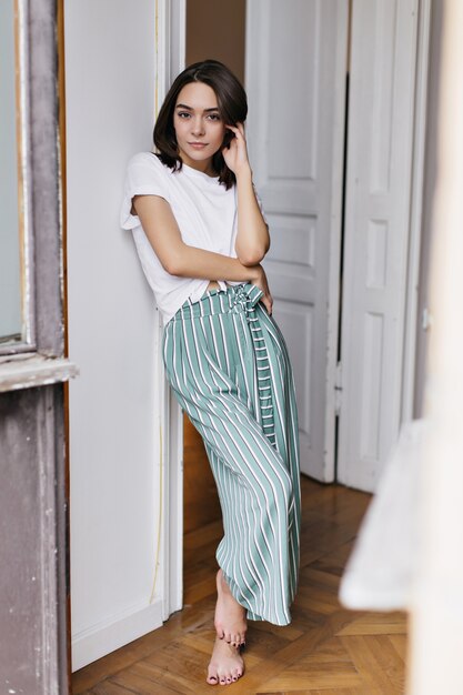 Barefooted young lady in cozy clothes posing at home. Brunette elegant girl in white t-shirt chilling in weekend in her apartment.