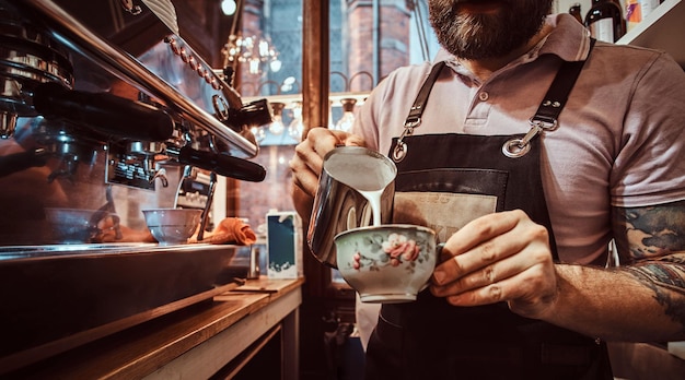 Barista in apron making a cappuccino, pouring milk in a cup in a restaurant or coffee shop