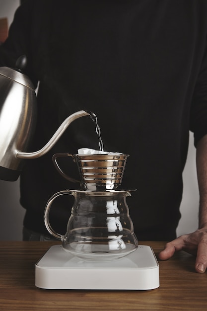 Free photo barista spills hot water to prepare filtered coffee from modern teapot to beautiful transparent drip coffee maker on white simple weights.