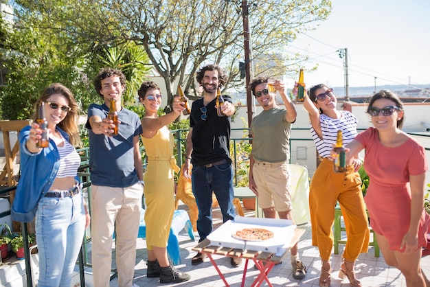 Free photo beaming friends clinking beer bottles. people of different nationalities raising bottles at camera, laughing on sunny day. party, friendship concept