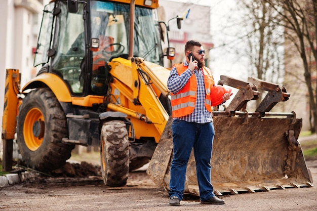 Free photo beard worker man suit construction worker in safety orange helmet sunglasses against tractor with mobile phone at hand