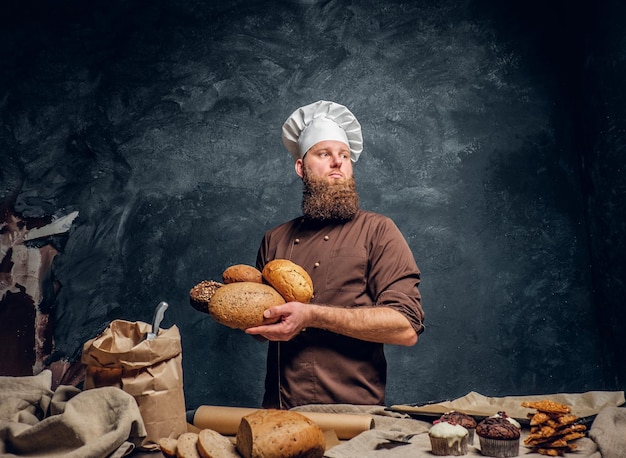 Free photo a bearded baker wearing a uniform holding fresh bread, standing next to a table, decorated with delicious bread loaves, baguettes and muffins in a dark studio