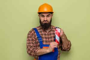 Free photo bearded builder man in construction uniform and safety helmet holding adhesive tape looking at camera confused standing over green background