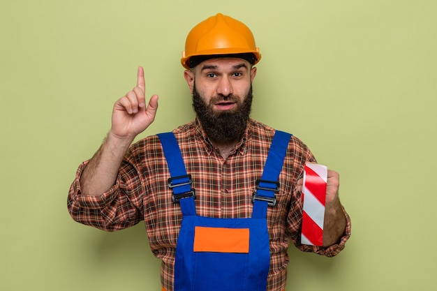 Free photo bearded builder man in construction uniform and safety helmet holding adhesive tape looking with smile on face showing index finger having new idea