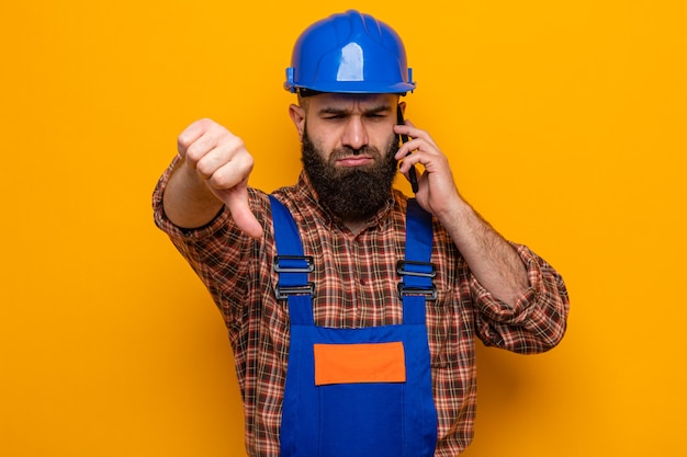 Free Photo bearded builder man in construction uniform and safety helmet looking at camera displeased showing thumbs down talking on mobile phone standing over orange background