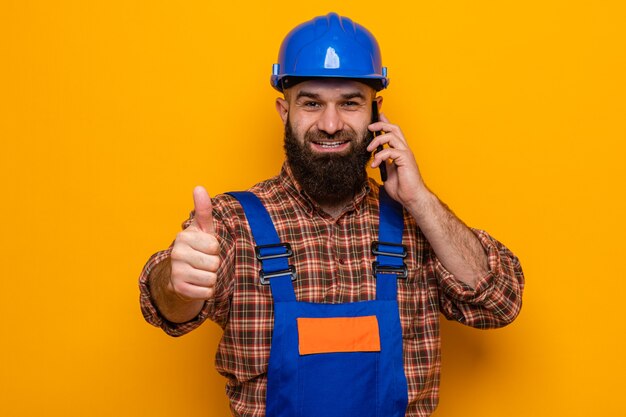 Bearded builder man in construction uniform and safety helmet looking smiling cheerfully showing thumbs up