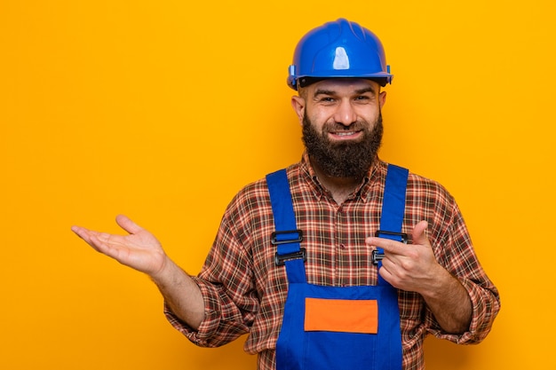 Free photo bearded builder man in construction uniform and safety helmet  smiling cheerfully presenting something with arm of his hand pointing with index finger to the side