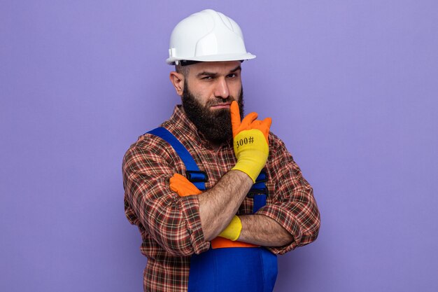 Bearded builder man in construction uniform and safety helmet wearing rubber gloves looking with serious face thinking