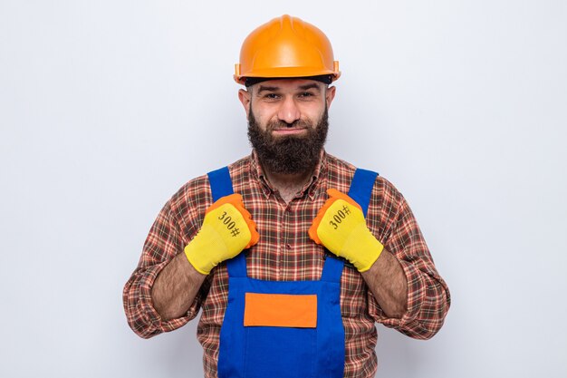 Bearded builder man in construction uniform and safety helmet wearing rubber gloves looking with smile on happy face