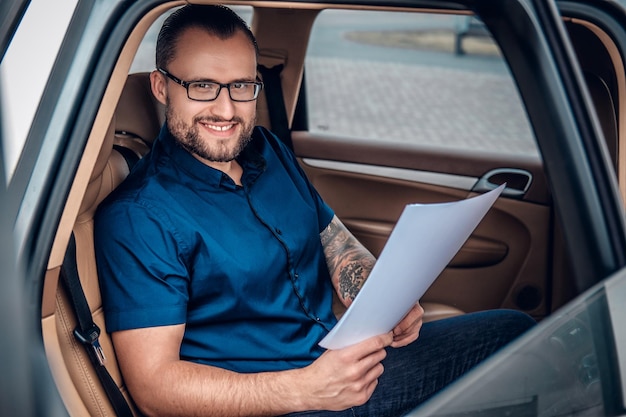 Free photo bearded business male in eyeglasses with tattoo on his arm sits on a back seat of a car.