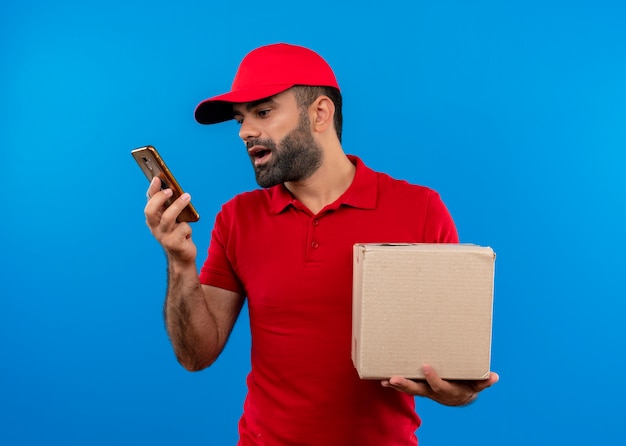 Bearded delivery man in red uniform and cap holding box package recording voice message using his smartphone standing over blue wall