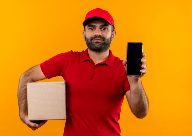 Bearded delivery man in red uniform and cap holding box package showing smartphone looking confident standing over orange wall