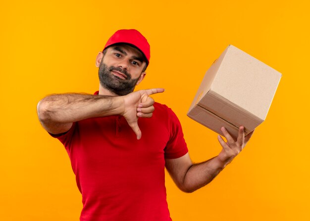 Bearded delivery man in red uniform and cap holding box package showing thumbs down standing over orange wall