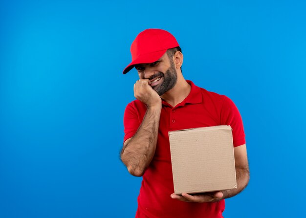 Bearded delivery man in red uniform and cap holding box package smiling happy and positive standing over blue wall