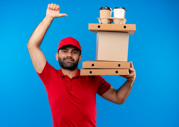 Bearded delivery man in red uniform and cap holding cardboard boxes  with smile on face showing thumbs up standing over blue wall