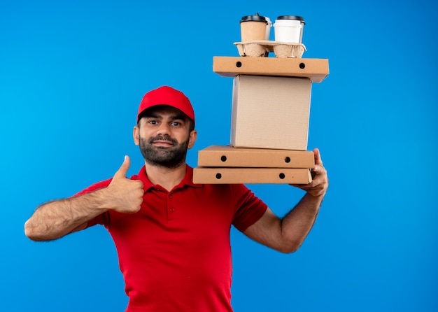 Bearded delivery man in red uniform and cap holding cardboard boxes  with smile on face showing thumbs up standing over blue wall