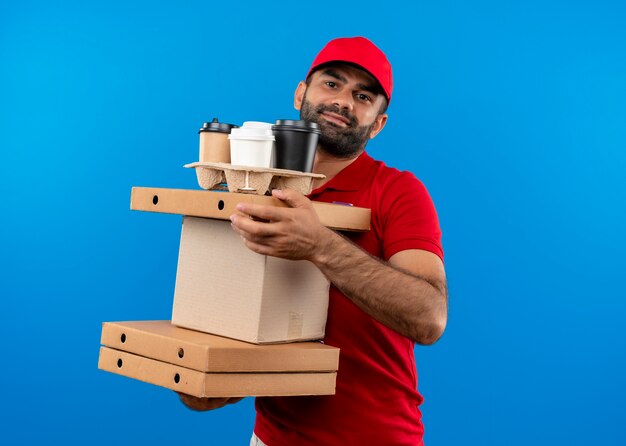 Bearded delivery man in red uniform and cap holding cardboard boxes  with smile on face standing over blue wall