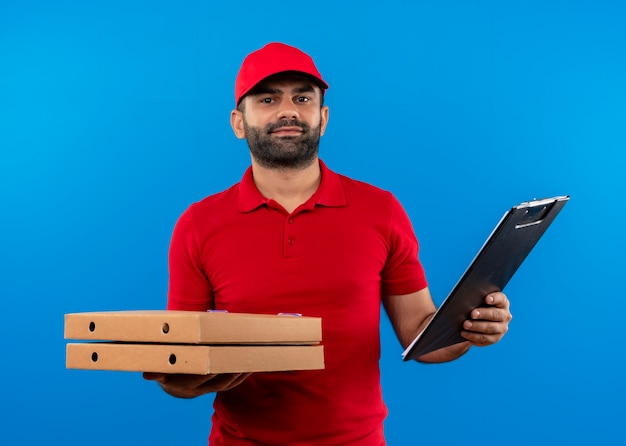 Bearded delivery man in red uniform and cap holding stack of pizza boxes and clipboard  with confident expression standing over blue wall