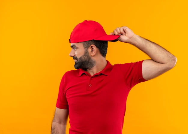 Bearded delivery man in red uniform and cap looking aside smiling turned his cap standing over orange wall