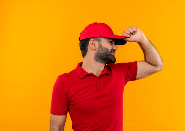 Bearded delivery man in red uniform and cap looking aside with hand on his cap lookin for someone standing over orange wall