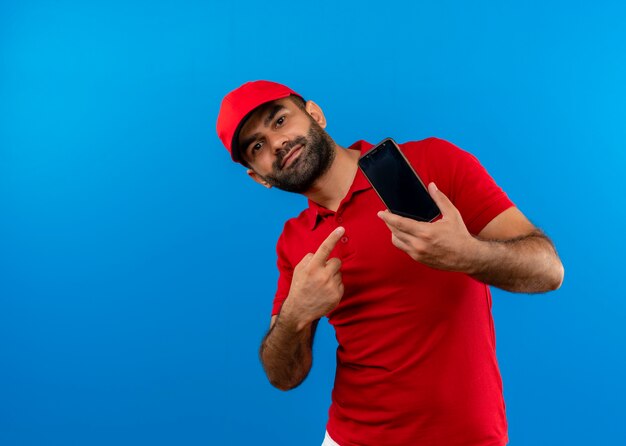 Bearded delivery man in red uniform and cap showing his smartphone pointing with finger to it smiling standing over blue wall