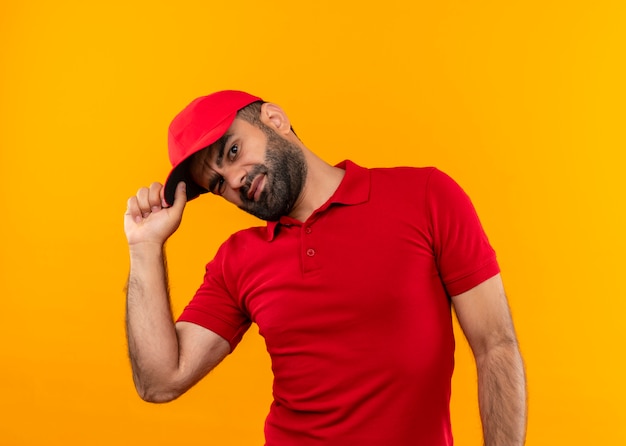 Free photo bearded delivery man in red uniform and cap  smiling and winking standing over orange wall