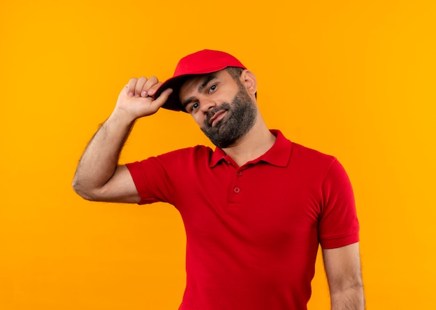 Bearded delivery man in red uniform and cap  with smile on face touching his cap standing over orange wall