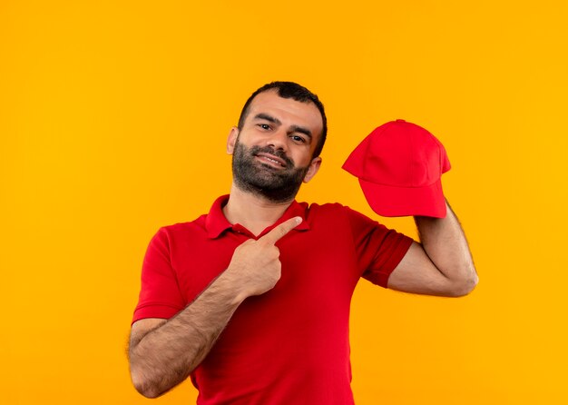 Bearded delivery man in red uniform holding cap pointing with finger to it smiling confident standing over orange wall