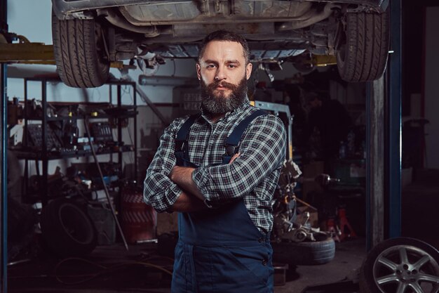 Bearded expert mechanic dressed in a uniform, standing with crossed arms against a car on a lift in the garage.