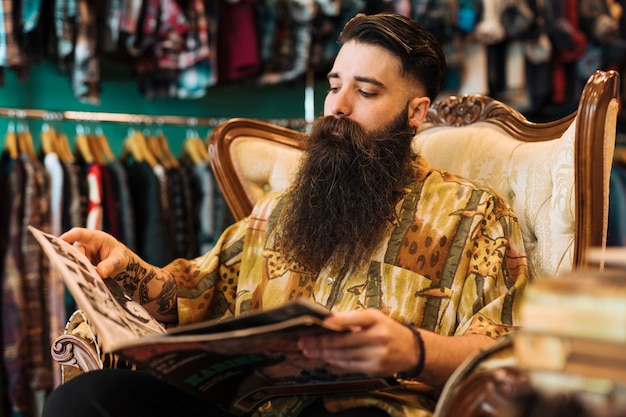 Bearded fashionable young man sitting on chair looking at magazine in the shop